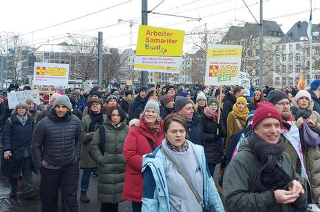 Die Mitarbeitenden des ASB NRW laufen mit drei Schildern Arbeiter-Samariter-Bunt" inmitten anderer Demonstranten zum Versammlungsplatz an der Deutzer Werft. 